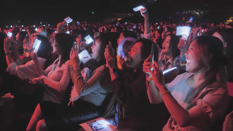 Bei einem K-Pop-Konzert sitzen reihenweise lächelnde Fans und filmen mit Handykameras.