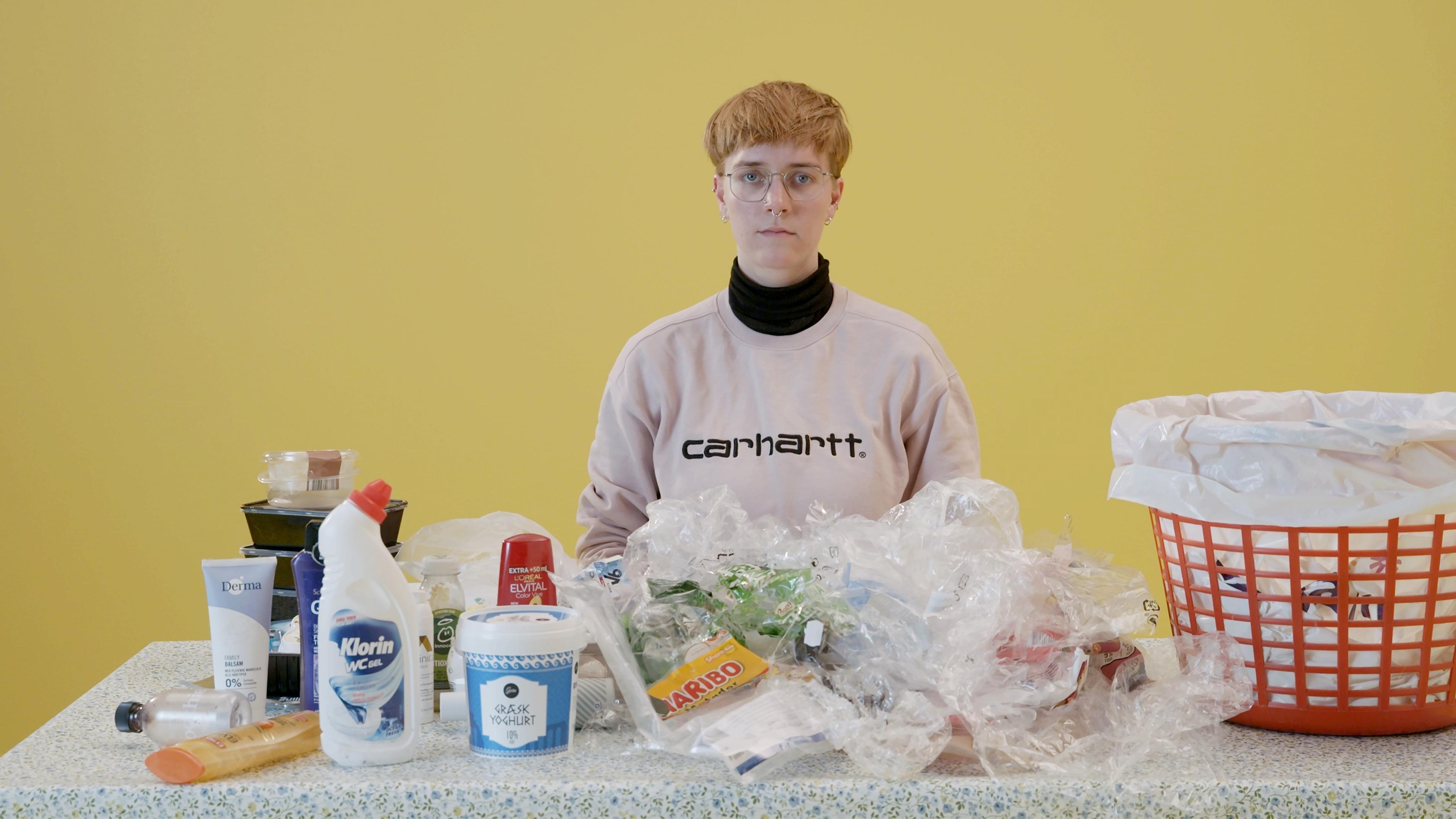 Eine junge Frau sitzt an einem Tisch, auf dem sich verschiedene Verpackungen aus Plastik befinden. Auf ihrer linken Seite steht ein großer Mülleimer.