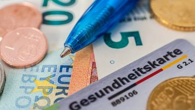 Eine Karte der Krankenversicherung liegt zusammen mit einem Kugelschreiber auf Geldscheinen und Münzen. 