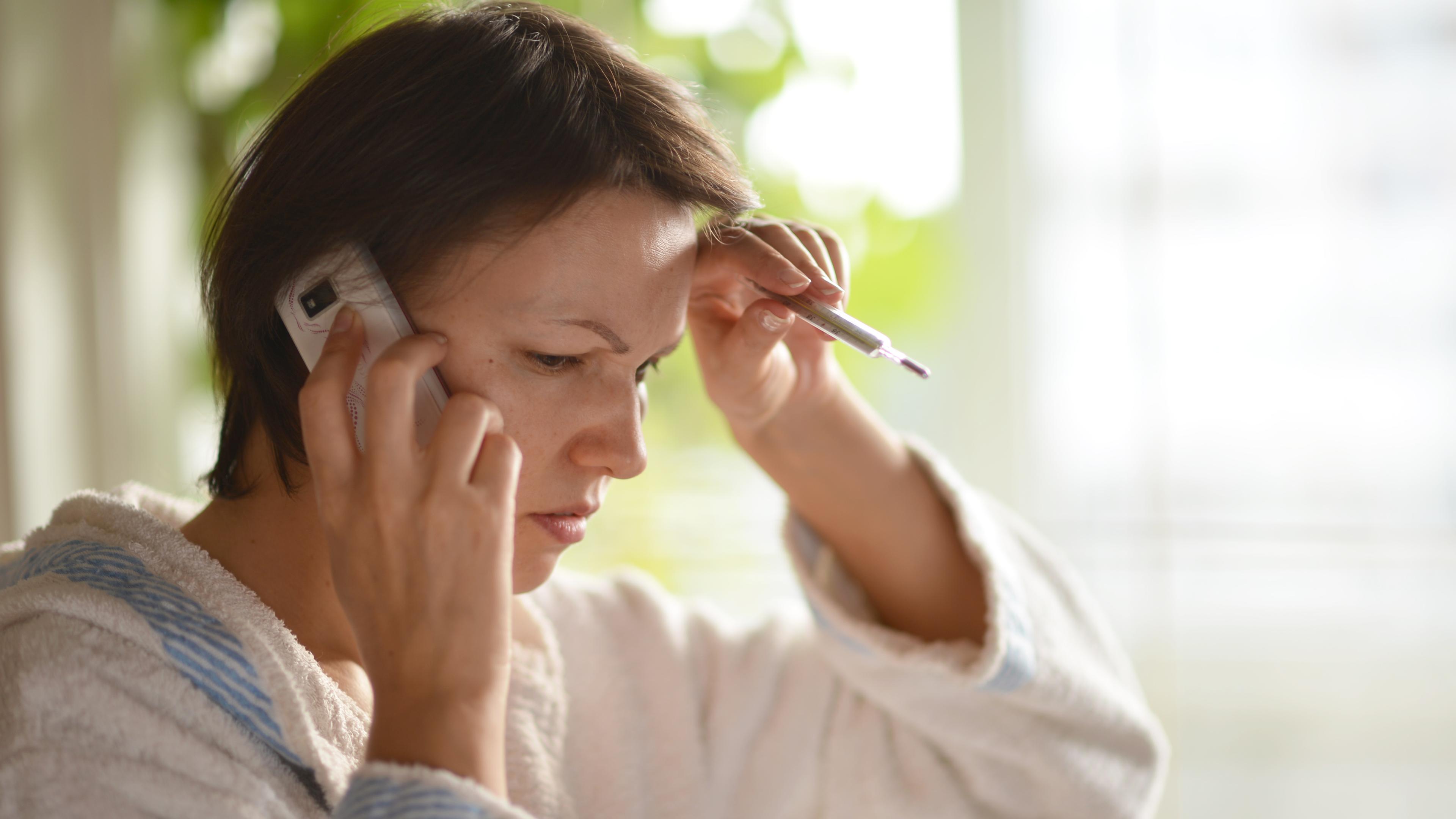Eine Frau mit Fieberthermometer in der Hand telefoniert.