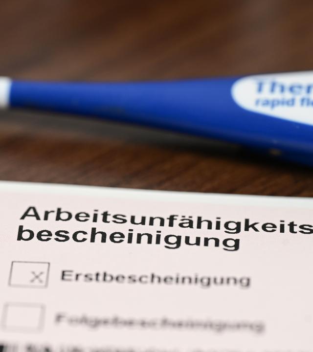 Ein Fieberthermometer, Medikamente und eine ärztliche Arbeitsunfähigkeitsbescheinigung (Krankmeldung) liegen auf einem Nachttisch