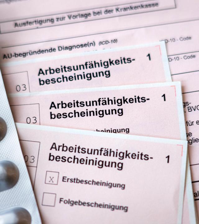 Mehrere Formulare zur Arbeitsunfähigkeitsbescheinigung liegen neben einem Blister mit Tabletten auf einem Tisch.