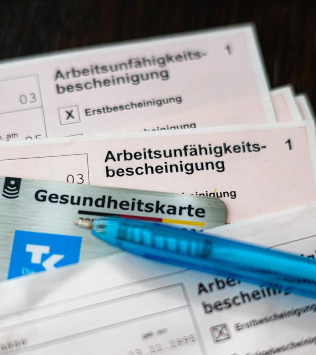 Eine Arbeitsunfähigkeitsbescheinigung liegt zusammen mit einem Kugelschreiber und einer elektronischen Gesundheitskarte auf einem Tisch.