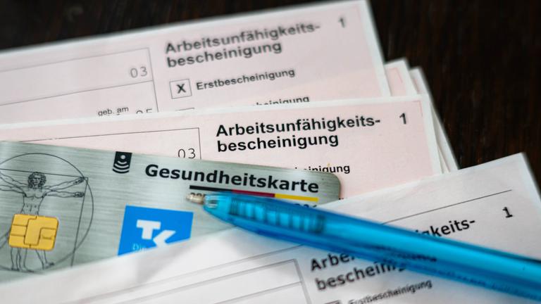 Eine Arbeitsunfähigkeitsbescheinigung liegt zusammen mit einem Kugelschreiber und einer elektronischen Gesundheitskarte auf einem Tisch.
