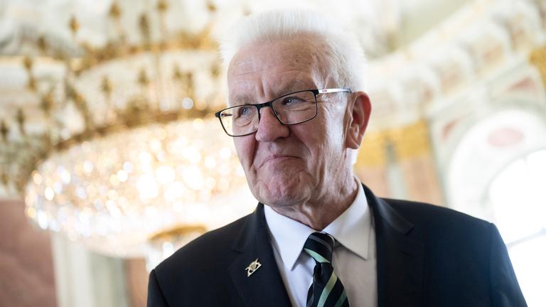 Winfried Kretschmann steht bei seiner Verabschiedung als Ministerpräsident im Neuen Schloss in Stuttgart, im Hintergrund ein funkelnder Kronleuchter.