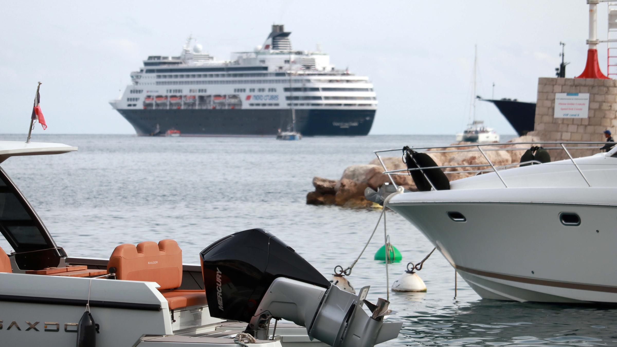 Ein Kreuzfahrtschiff ist am Horizont zu sehen. Im Vordergrund sieht man ankernde kleinere Schiffe im Hafen.