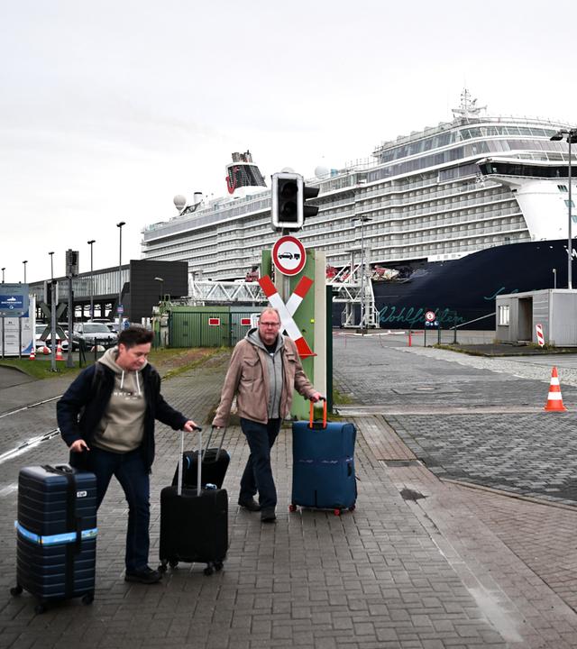Passagiere der "Mein Schiff 3" verlassen mit Gepäck das Kreuzfahrtterminal, hinten steht das Schiff.