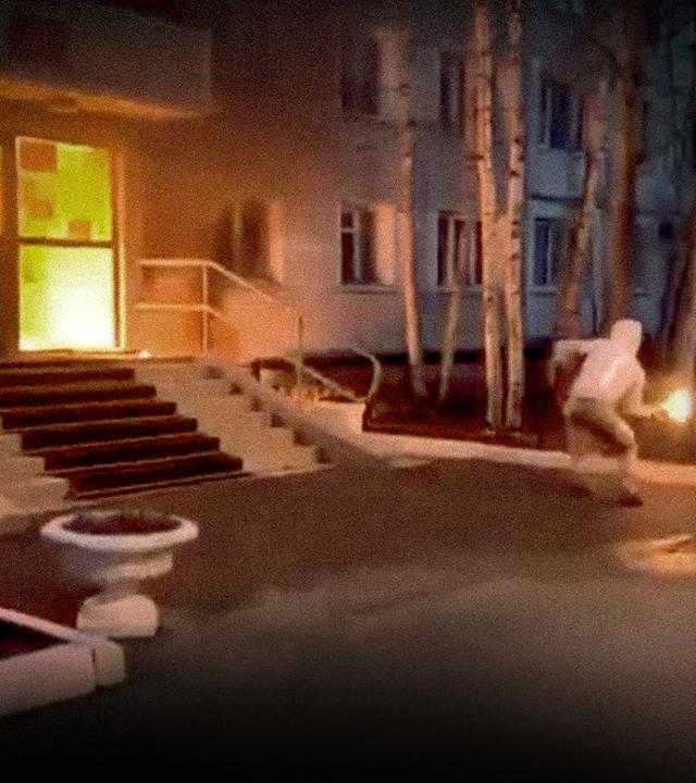 Links, ein brennendes Gebäude, mitte/rechts, eine vermummte Person die einen Molotow-Cocktail auf das brennende Gebäud wirft.