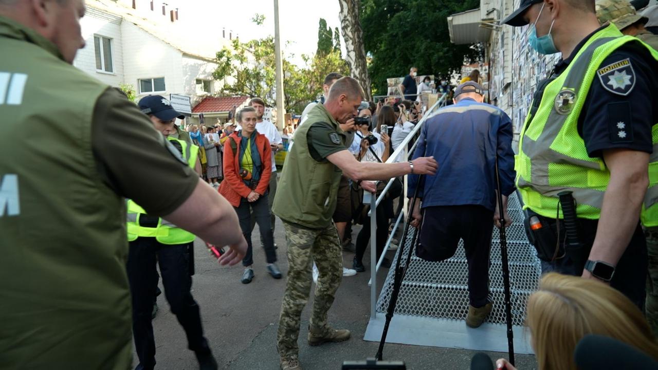 Ein Mann läuft auf Krücken in das Gebäude. Ihm fehlt ein Bein und er wird begeleitet von einem Soldaten.
