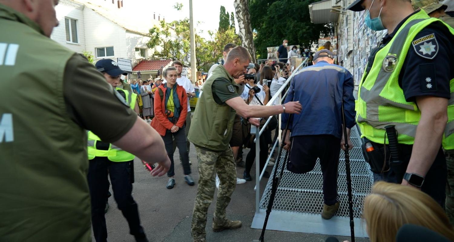 Ein Mann läuft auf Krücken in das Gebäude. Ihm fehlt ein Bein und er wird begeleitet von einem Soldaten.