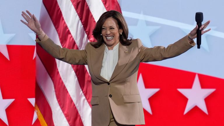Kamala Harris mit ausgebreiteten Armen und Mikro in der rechten Hand vor einer amerikanischen Flagge