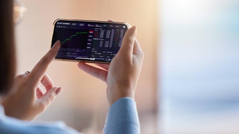 Eine Frau hält ein Smartphone in der Hand, auf dem die Kurve einer Kursentwicklung zu sehen ist.