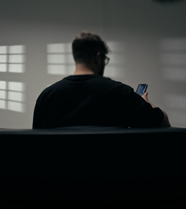 Ein Mann von hinten sitzt auf einem Sofa und schaut auf sein Handy. An der Wand reflektieren sich die Umrisse der Fenster.