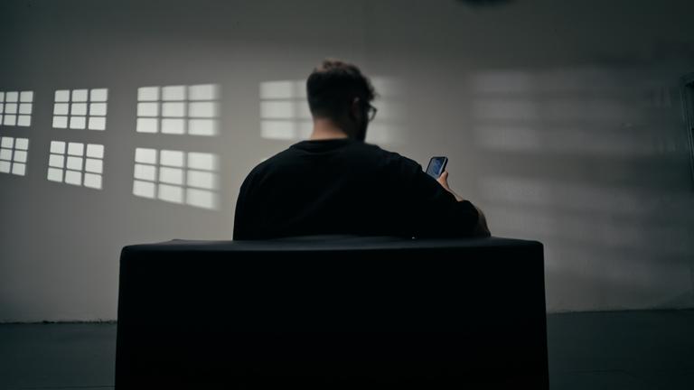 Ein Mann von hinten sitzt auf einem Sofa und schaut auf sein Handy. An der Wand reflektieren sich die Umrisse der Fenster.