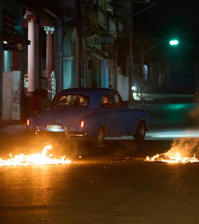 13.05.2026, Kuba, Havanna: Menschen fahren mit einem amerikanischen Oldtimer durch eine Barrikade, die von Anwohnern errichtet wurde, die gegen anhaltende Stromausfälle in Havanna protestieren.