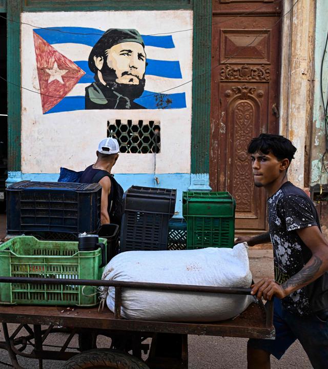 Ein junger Mann schiebt ein Lastenrad, im Hintergrund eine Wandbemalung von Fidel Castros Gesicht auf der kubanischen Flagge