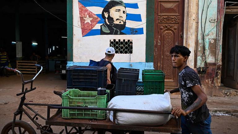 Ein junger Mann schiebt ein Lastenrad, im Hintergrund eine Wandbemalung von Fidel Castros Gesicht auf der kubanischen Flagge