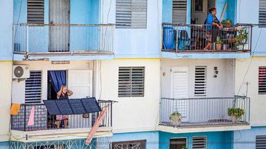 ein Ausschnitt von einem blauen Wohnhaus mit Balkonen. Auf zwei Balkonenen steht jeweils eine Frau. Vor dem Balkon der einen Frau sind improvisierte Solar Panels angebracht.