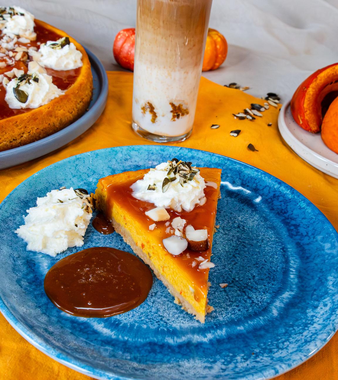 Ein Stück Kürbis-Cheesecake auf einem blauen Teller. Im Hintergrund sind der ganze Cheesecake, ein Getränk und drei Scheiben Kürbis zu sehen.