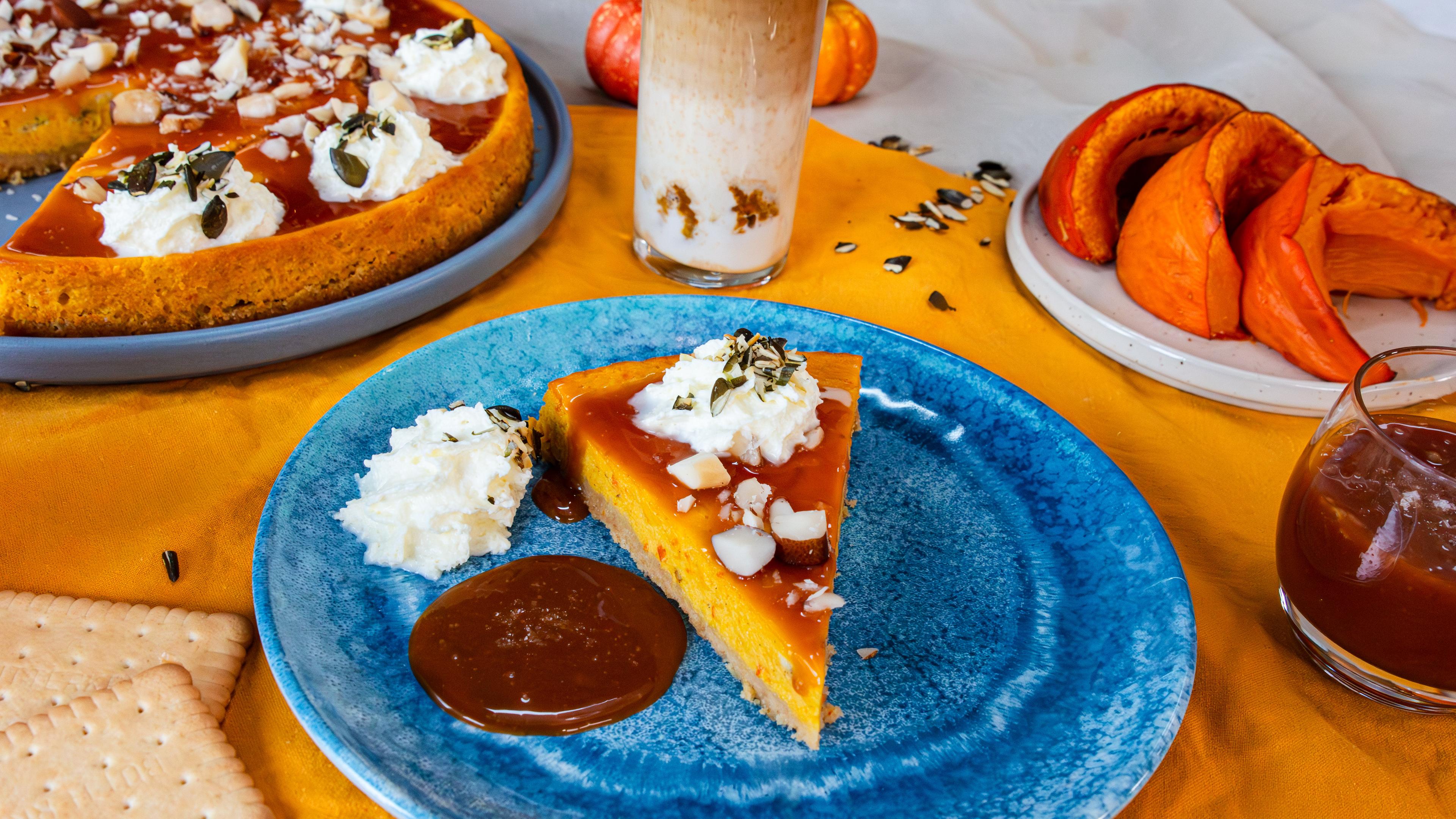 Ein Stück Kürbis-Cheesecake auf einem blauen Teller. Im Hintergrund sind der ganze Cheesecake, ein Getränk und drei Scheiben Kürbis zu sehen.