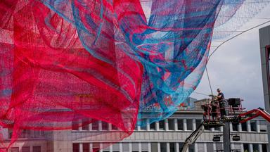 Das Bild zeigt die große Kunstinstallation "A Sky Full of Hope", die aus blauen und roten Fäden besteht, die zwischen hohen Gebäuden gespannt sind.