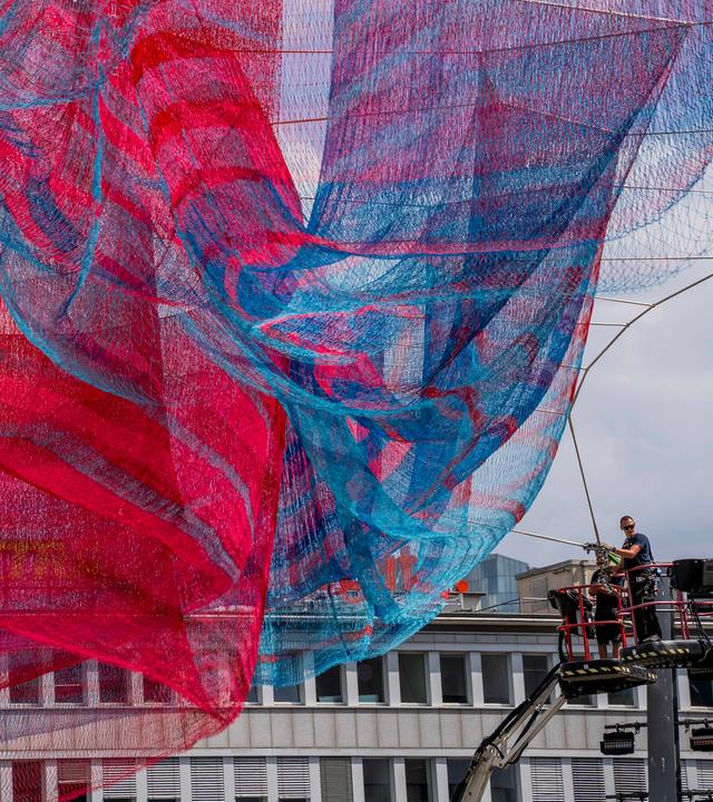 Das Bild zeigt die große Kunstinstallation "A Sky Full of Hope", die aus blauen und roten Fäden besteht, die zwischen hohen Gebäuden gespannt sind.