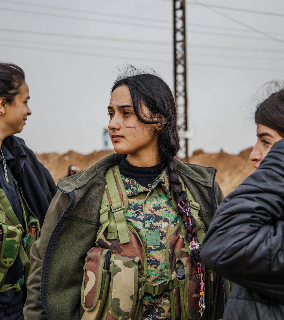 Drei junge Frauen mit dunklen Haaren tragen dicke schwarze Winterjacken über militärischen Camouflage-Westen, stehen nebeneinander und schauen in verschiedene Richtungen.