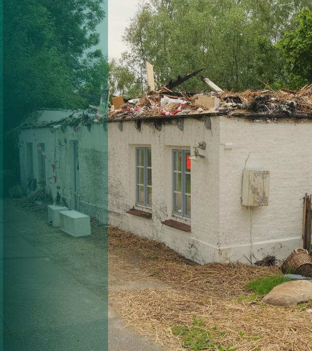 Ein weiß-gestrichenes Haus am Waldrand, die obere Etage fehlt, dort sind nur noch verbrannte Überreste sichtbar.
