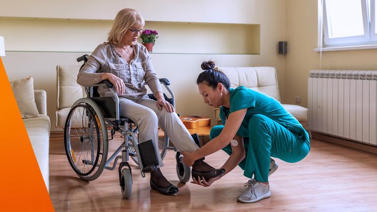 Pflegerin zieht einer Frau im Rollstuhl einen Schuh an