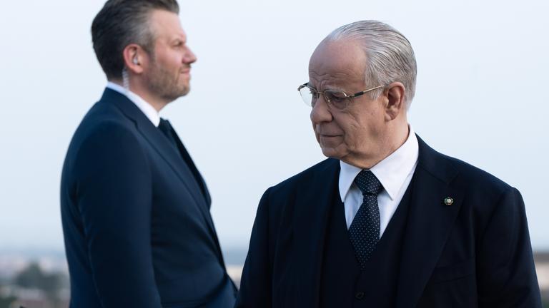 Im Vordergrund steht ein älterer Mann im Anzug mit Brille, der nachdenklich zur Seite schaut. Hinter ihm steht sein Bodyguard, ein Mann mittleren Alters, ebenfalls im Anzug und Knopf im Ohr.