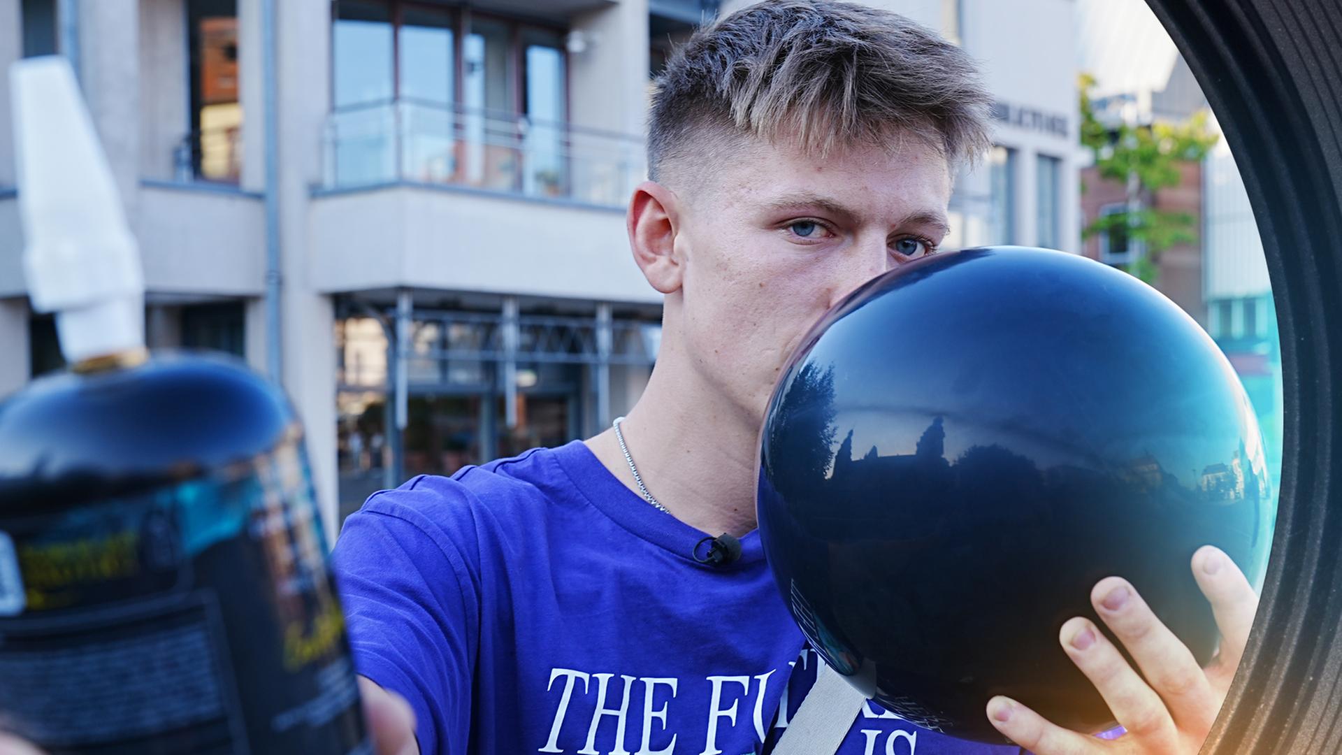 Ein junger Mann hält sich mit einer Hand einen Luftballon vor den Mund. Mit der anderen Hand streckt er eine Kartusche Lachgas in die Kamera.