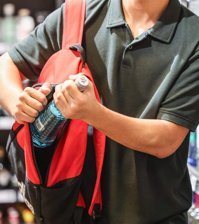 Ein Mann steckt in einem Supermarkt eine Flasche mit einem alkoholhaltigen Getränk in seinen Rucksack. 
