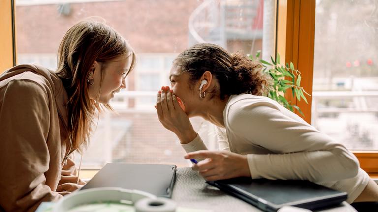 Seitenansicht eines weiblichen Teenagers, der mit einer Freundin an einem Tisch sitzt und tratscht.