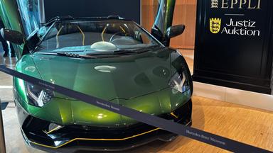 Auf dem Bild ist zu sehen: Ein metallicgrüner Lamborghini Aventador Roadster mit nach oben offenen Türen. Rechts neben dem Wagen steht eine schwarze Tafel mit den Aufschriften „Eppli”, „Justiz-Auktion” und dem baden-württembergischen Landeswappen.