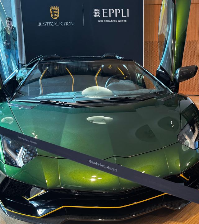 Auf dem Bild ist zu sehen: Ein metallicgrüner Lamborghini Aventador Roadster mit nach oben offenen Türen. Rechts neben dem Wagen steht eine schwarze Tafel mit den Aufschriften „Eppli”, „Justiz-Auktion” und dem baden-württembergischen Landeswappen.