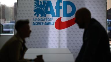 Landesparteitag der AfD Sachsen-Anhalt im April 2026: Zwei Teilnehmende stehen vor dem Parteilogo