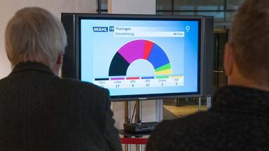 Zwei SPD-Mitglieder in Wlly-Brandt-Haus sitzen vor Fernsehbildschirm, der die Sitzverteilung im Thüringer Landtag nach der ersten Hochrechnung abbildet