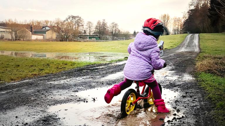Kind fährt mit dem Laufrad auf einem matschigen Feldweg durch die Pfütze