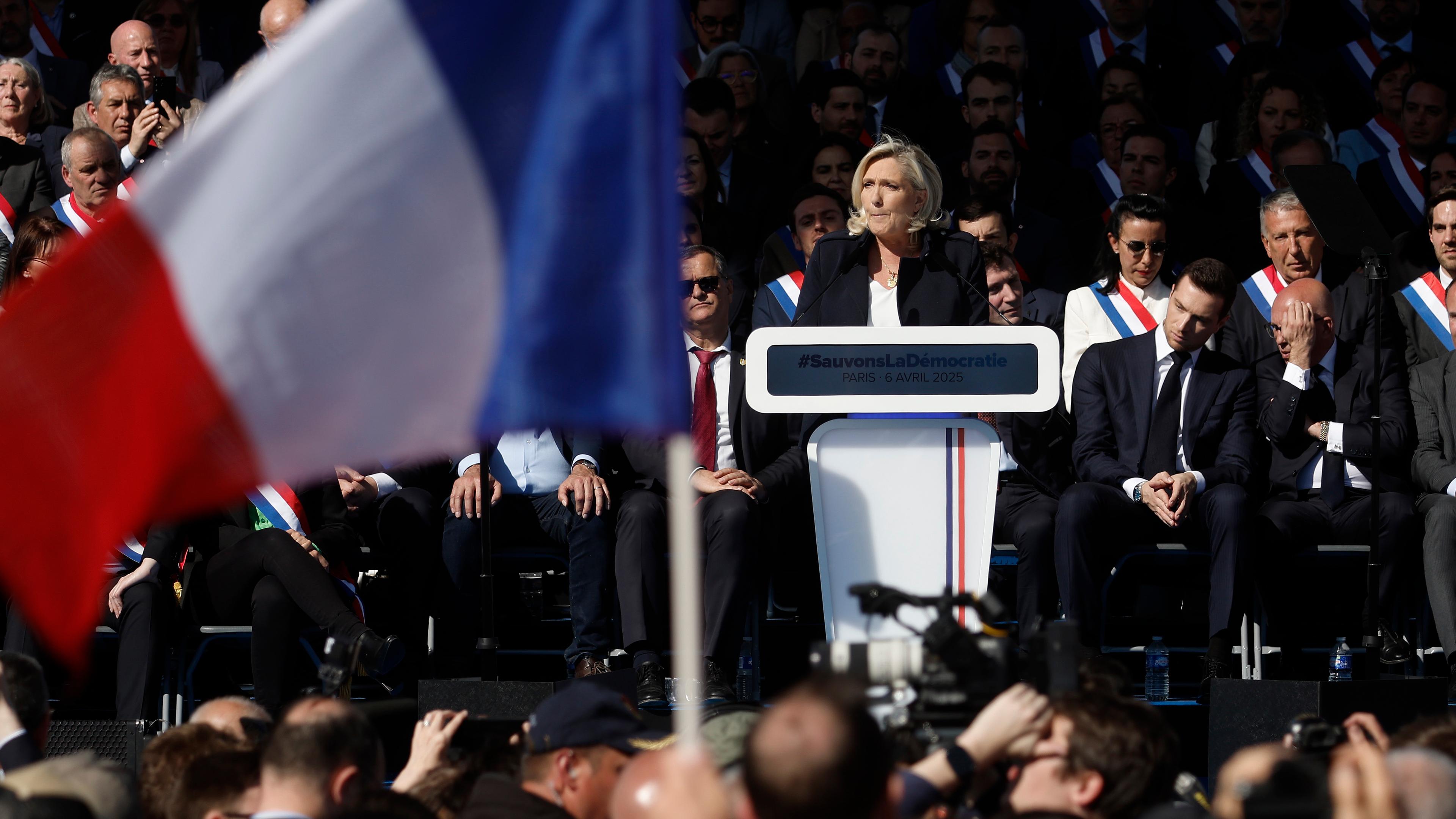 Marine Le Pen steht an einem Rednerpult, im Vordergrund weht eine französische Flagge.