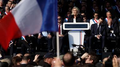 Marine Le Pen steht an einem Rednerpult, im Vordergrund weht eine französische Flagge.
