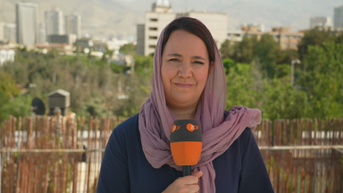 Auf dem Bild ist Phoebe Gaa in Teheran zu sehen. Auf dem Bild hält Sie ein Mikrofon und im Hintergrund sind Fahrzeuge unterwegs.