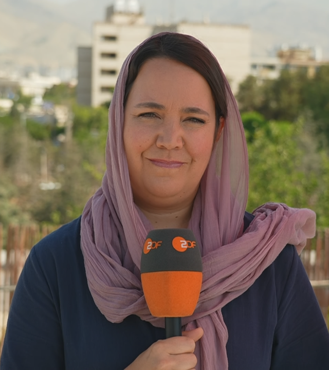 Auf dem Bild ist Phoebe Gaa in Teheran zu sehen. Auf dem Bild hält Sie ein Mikrofon und im Hintergrund sind Fahrzeuge unterwegs.