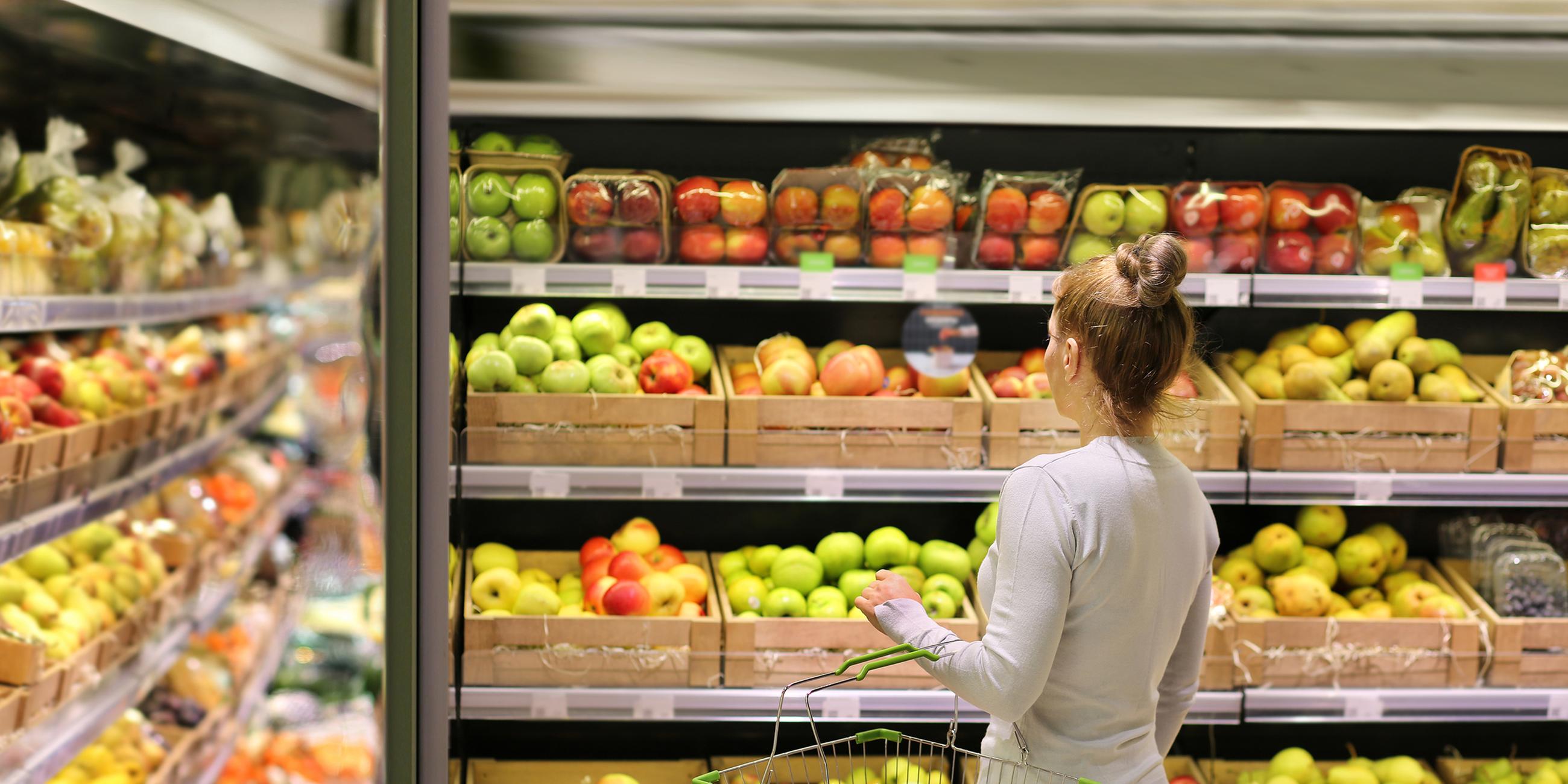 Eine Frau steht mit einem kleinen Einkaufskorb vor dem Obstregal im Supermarkt.