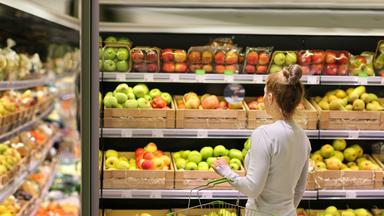 Eine Frau steht mit einem kleinen Einkaufskorb vor dem Obstregal im Supermarkt.