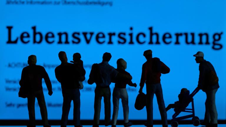 Modellfiguren stehen vor einem blau leuchtendem Display mit der Aufschrift "Lebensversicherung".