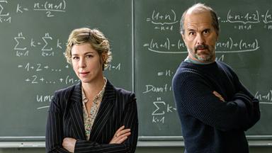 "Lehrer kann jeder!": Richard Glossat (Christoph Maria Herbst) sitzt in einem Klassenzimmer auf dem Lehrerpult und hält ein Mathematik-Buch in der Hand. Neben ihm steht Anke Glossat (Brigitte Zeh) mit verschränkten Armen und genervtem Blick. Im Hintergrund eine Schultafel, an die mit Kreide Mathematik-Formeln geschrieben stehen.