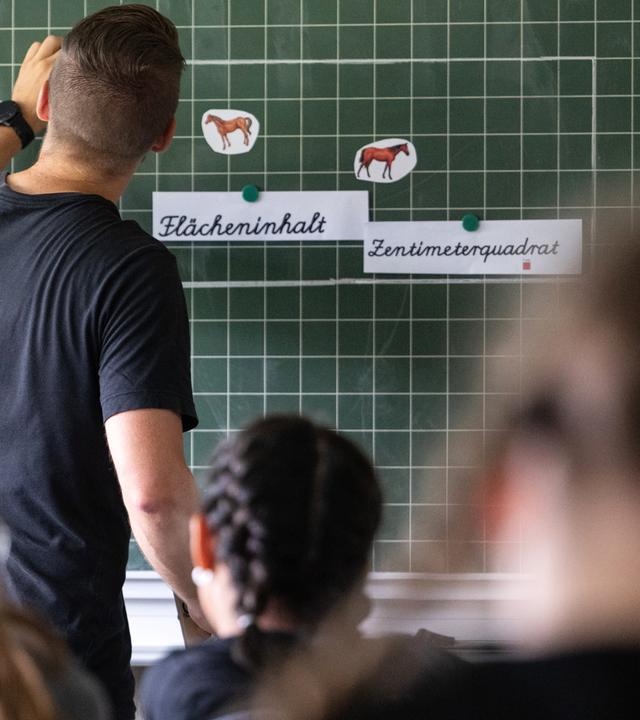 Ein Lehrer macht im Unterricht einer vierten Klasse einer Grundschule einen Tafelanschrieb, während an der Tafel Zettel mit der Aufschrift «Flächeninhalt» und «Zentimeterquadrat» hängen