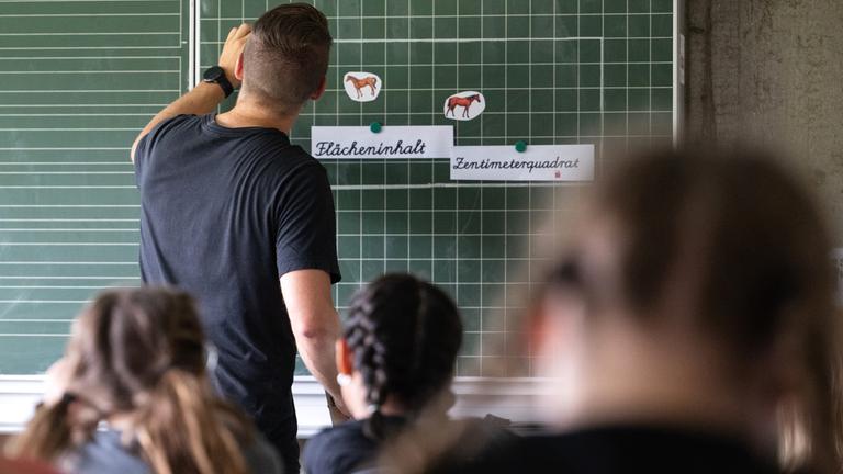 Ein Lehrer macht im Unterricht einer vierten Klasse einer Grundschule einen Tafelanschrieb, während an der Tafel Zettel mit der Aufschrift «Flächeninhalt» und «Zentimeterquadrat» hängen