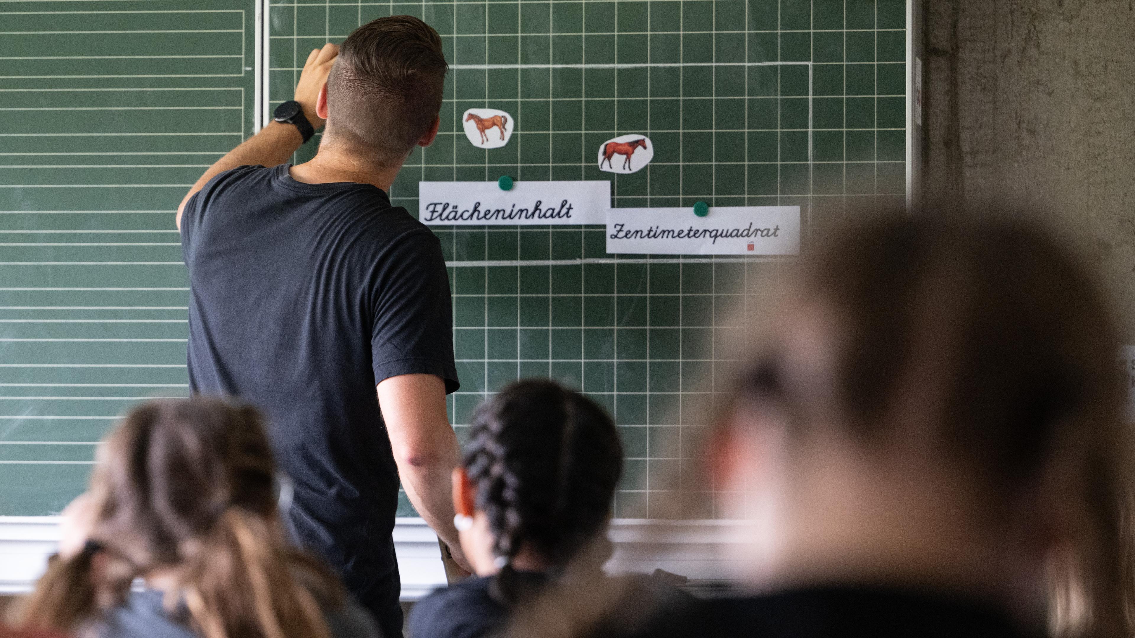 Ein Lehrer macht im Unterricht einer vierten Klasse einer Grundschule einen Tafelanschrieb, während an der Tafel Zettel mit der Aufschrift «Flächeninhalt» und «Zentimeterquadrat» hängen.