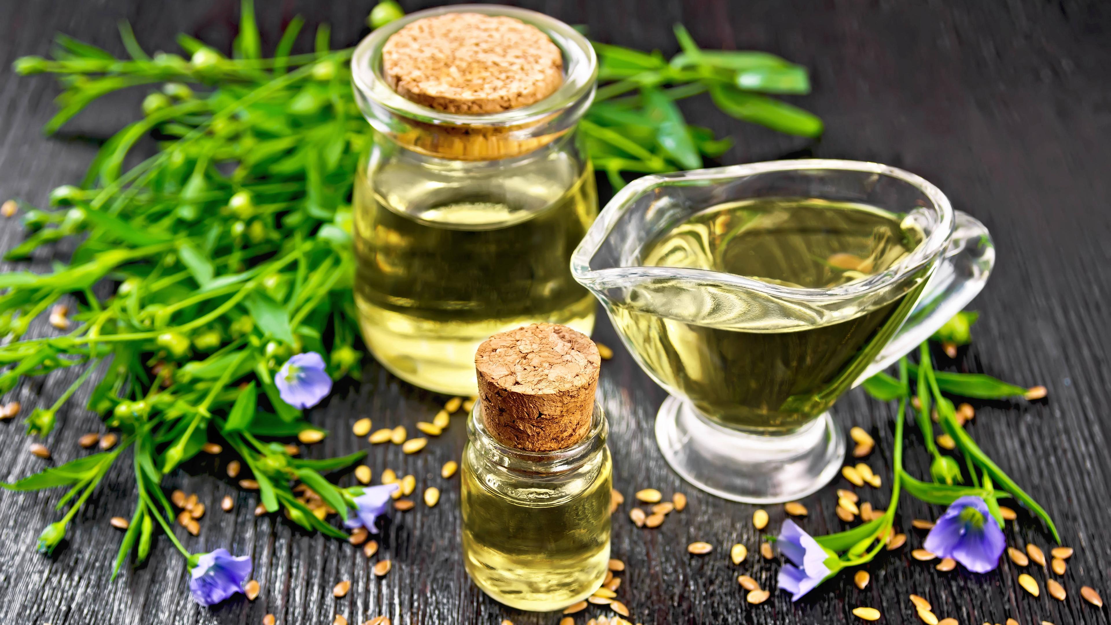 Auf einer schwarzen Oberfläche stehen drei kleine Glasbehälter, in denen Leinöl enthalten ist. Um die Behälter garniert liegen Flachsblüten und Leinsaat.